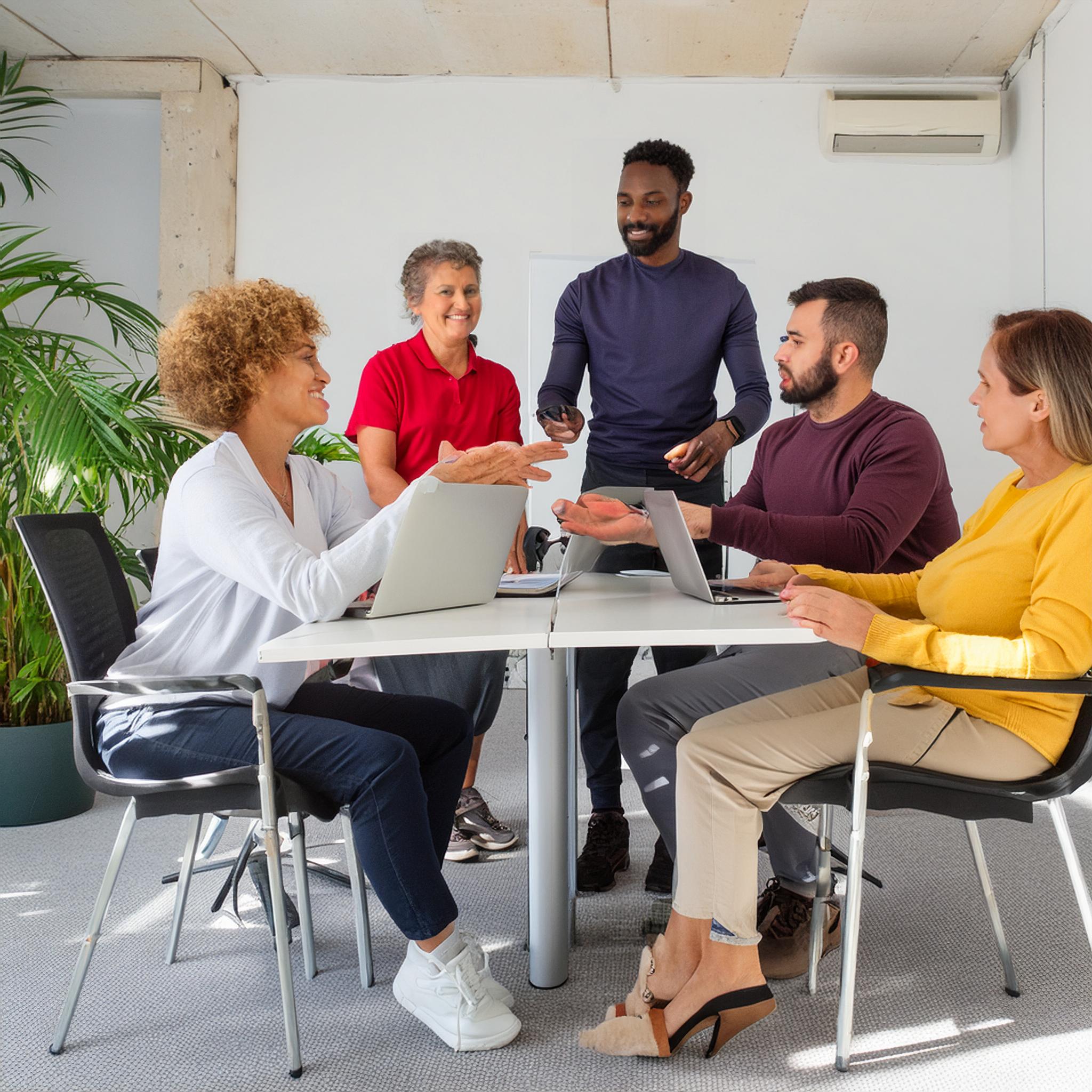 Menschen unterschiedlicher Herkunft und Altersklassen sitzen an einem Tisch mit Laptops und unterhalten sich